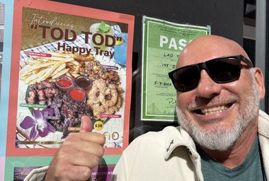 Tod Hirsch giving a thumbs up beside a Thai restaurant poster advertising a "Tod Tod" happy tray